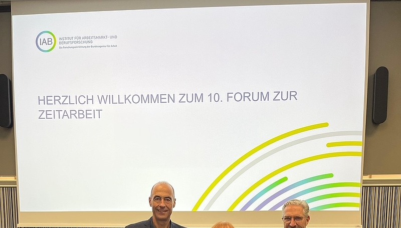 Ingrid Hofmann, Gründerin von Hofmann Personal, steht beim 10 Interdisziplinären Forum zur Zeitarbeit neben zwei Mitarbeitern in Anzügen.