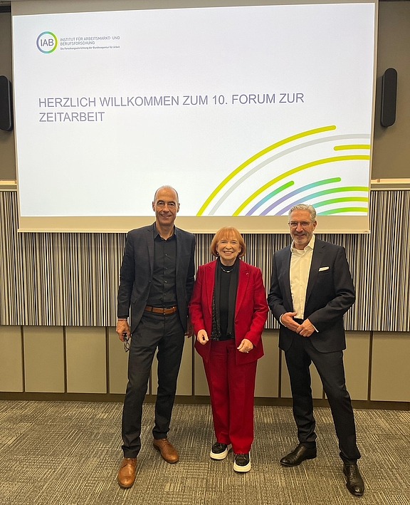 Ingrid Hofmann, Gründerin von Hofmann Personal, steht beim 10 Interdisziplinären Forum zur Zeitarbeit neben zwei Mitarbeitern in Anzügen.