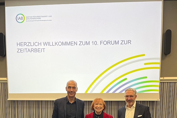 Ingrid Hofmann, Gründerin von Hofmann Personal, steht beim 10 Interdisziplinären Forum zur Zeitarbeit neben zwei Mitarbeitern in Anzügen.